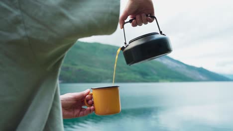 man pouring hot coffee into a cup with lake and mountain in the background