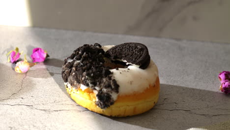 a hand with pink nails reaches to take a donut topped with crushed cookies and a whole oreo. set on a textured gray surface with scattered rose petals, the scene is warmly lit by natural daylight.