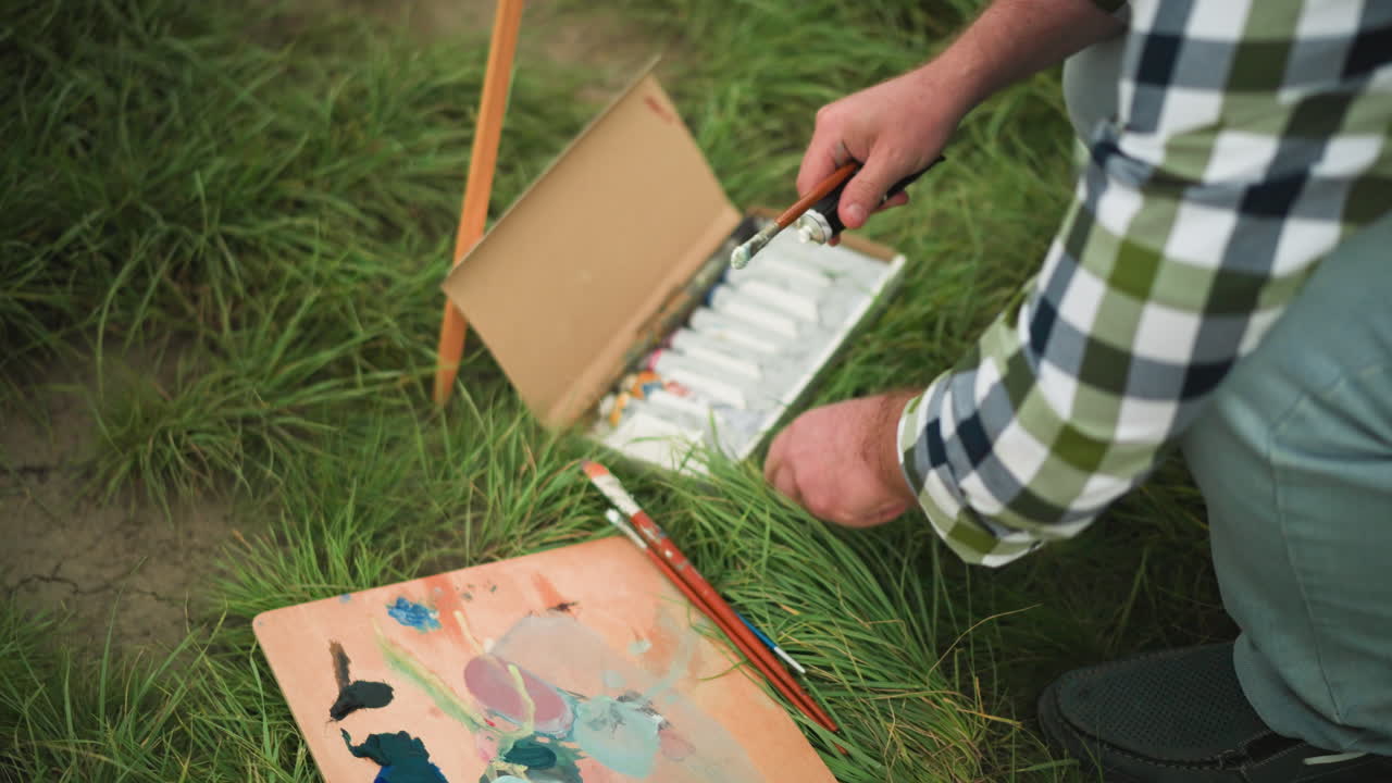 An artist in a checkered shirt bends down in a grass field, carefully opening a paint tube from a box of paints. The scene captures the preparation stage of outdoor painting