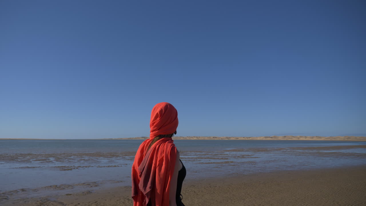 Kvinde med en rød turban, der går på det våde sand ved Naila-lagunen i Marokko. khenifiss nationalpark