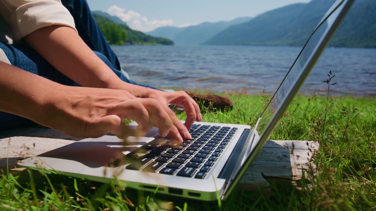 Person taster på bærbar computer ved siden af rolig sø, grønne bakker bagved. Hænder bevæger sig hurtigt, fokuseret på skærmen. Fredelig udendørs arbejdsscene, naturligt lys. Ideel til fjernarbejde, rejser eller digitale nomadetemaer.