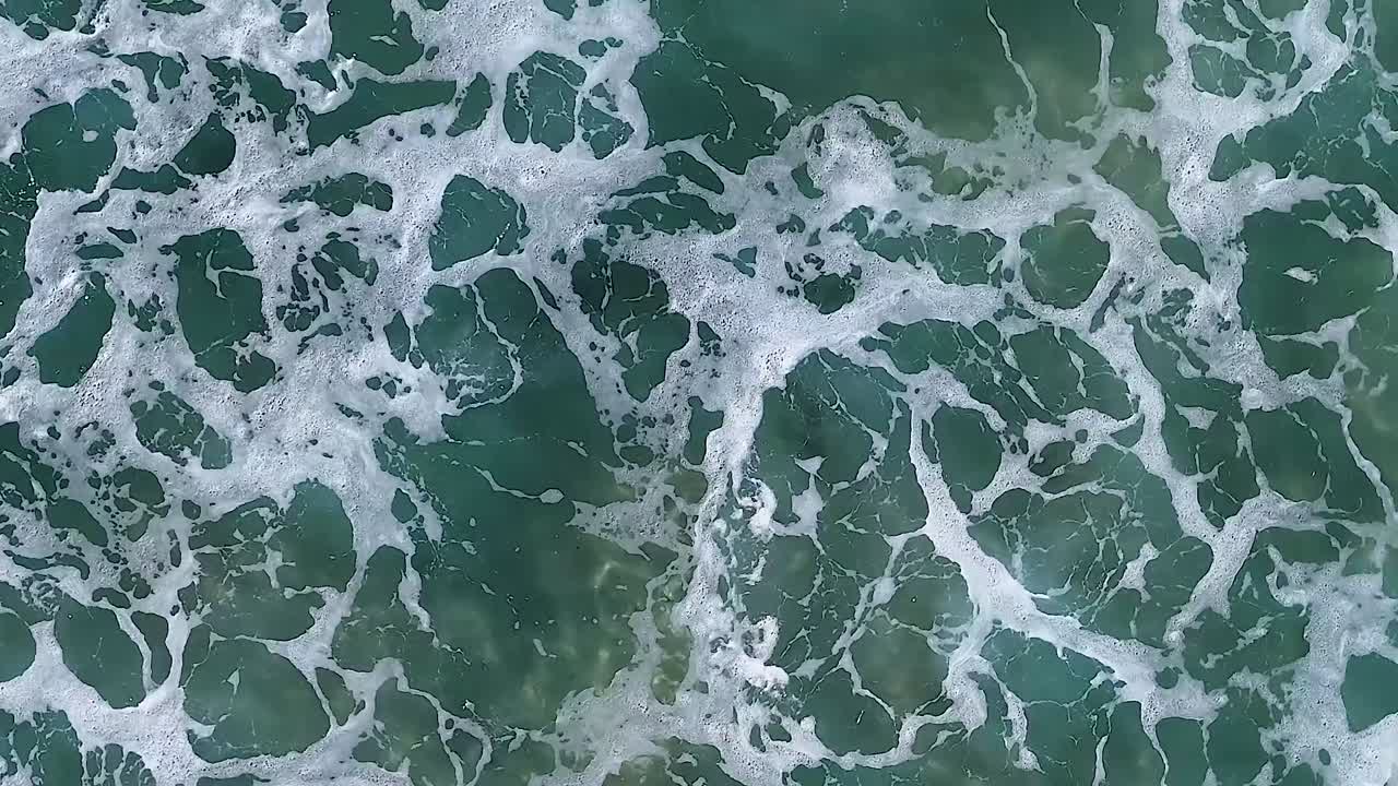 Aerial overhead shot of turbulent white sea foam swirling over deep teal ocean water