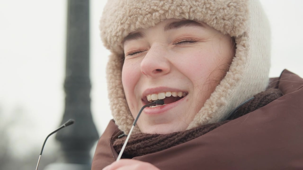 Close-up portrait of young person outdoors in cold overcast light, smiling with eyes closed and mouth slightly open while lowering eyeglasses by temple arm toward chin, wearing wool trapper hat and