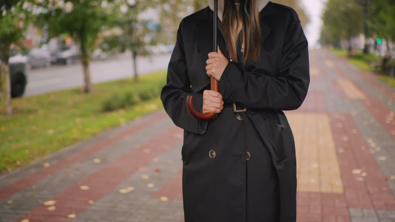 Close-up of woman in black trench coat holding umbrella with wooden handle standing on urban walkway during rainy autumn day, blurred background of cars, trees, pavement, and fallen leaves enhancing moody