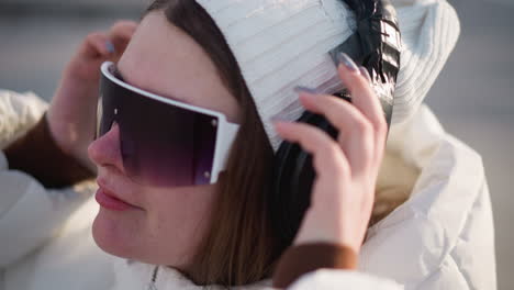 fair skinned girl with precise manicure places over ear headphones on ear, smiling lightly as she grooves to music in sunny winter urban street scene with gentle breeze and soft glow