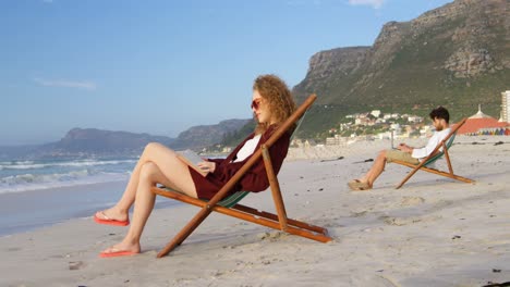 side view of young caucasian woman with sunglasses sitting on lounger and reading a book at beach 4k