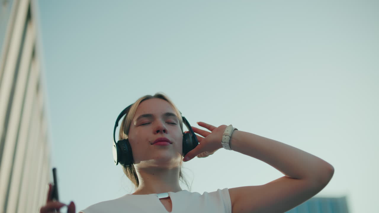 Upper angle view of music artist dancing to rhythm of song she is listening to with headphones on, hand raised, under clear sky with partial view of residential building