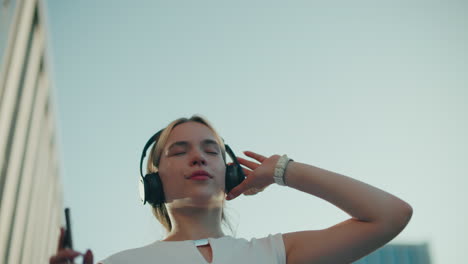 upper angle view of music artist dancing to rhythm of song she is listening to with headphones on, hand raised, under clear sky with partial view of residential building