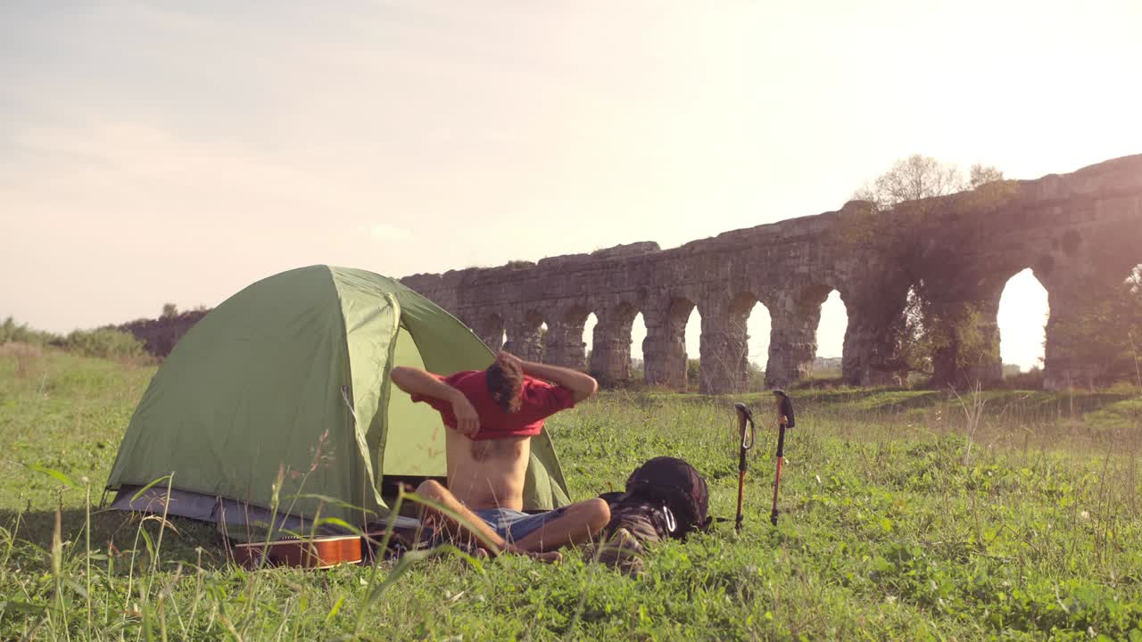 젊은 남자 배 타는 옷을 입고 티셔츠를 입고 캠핑 텐트 밖에서 잔디에 앉아 로마의 수로 앞에 파르코 데글리 아쿠에도티 공원 유적 로마에서 해가 지는 동안 땅에 서서히 움직이는 막대기
