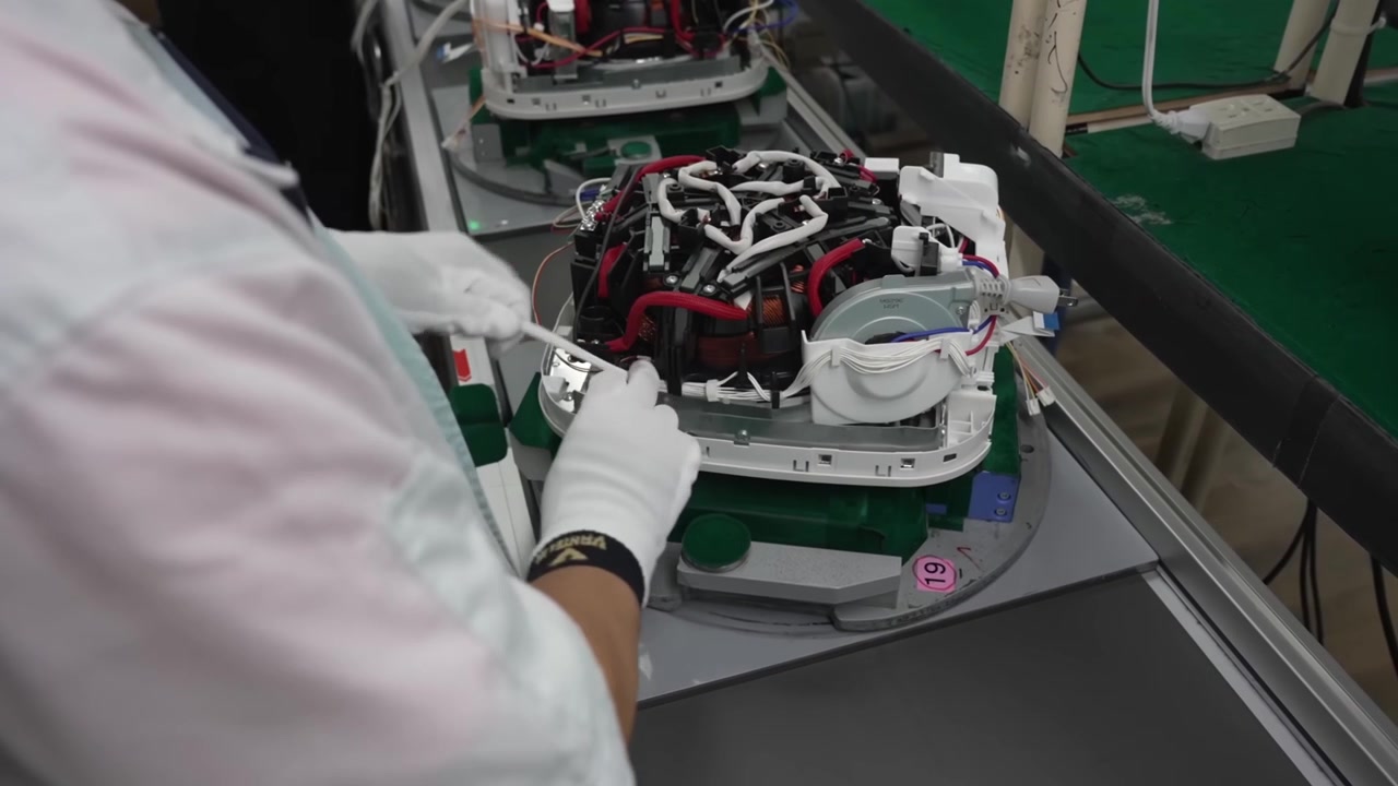 Worker handling partially assembled rice cooker unit with visible wiring on moving assembly conveyor belt