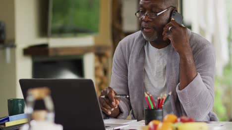 Nachdenklicher-Afroamerikanischer-älterer-Mann-Im-Bademantel,-Der-Einen-Laptop-Benutzt-Und-Auf-Dem-Smartphone-Spricht