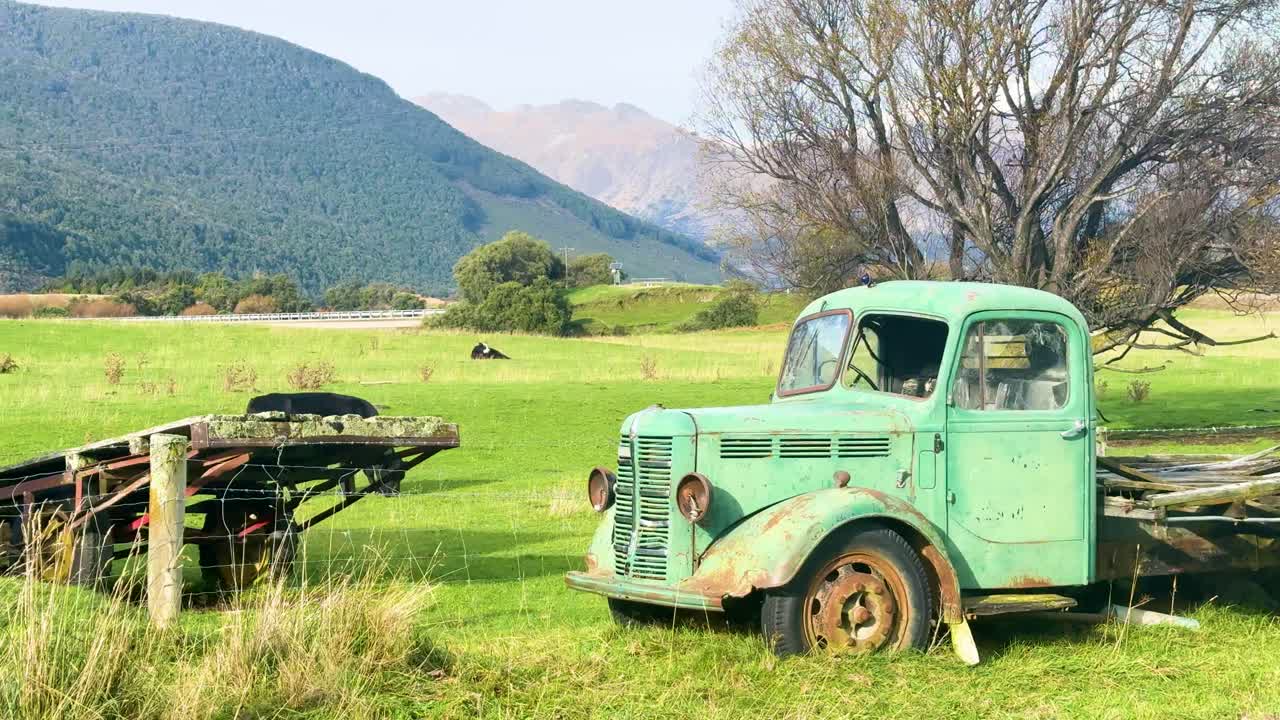 An old green truck sits in a tranquil field with mountains in the background, under bright daylight