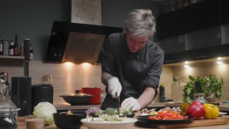 Young-professional-male-chef-in-an-elegant-black-shirt-with-an-alternative-look,-tattoos,-gloves-slicing-fresh-mushrooms-A-modern,-fancy-looking-kitchen-with-fresh-vegetables-besides-Cinematic-Sliding