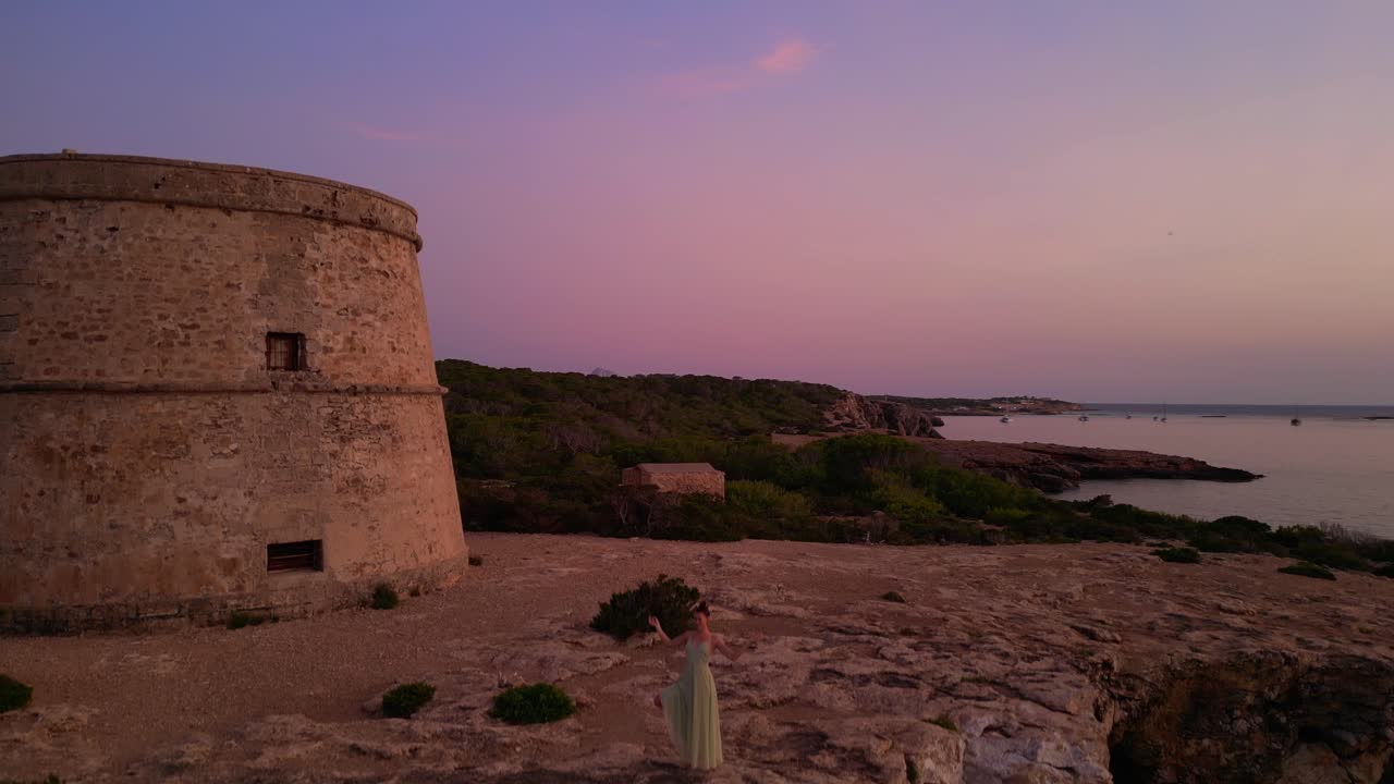 Woman performing yoga poses at sunset near Torre Rovira in Ibiza, Spain. Marvelous aerial view flight descending drone