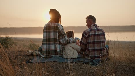 Rear-view-of-a-happy-brunette-girl-in-a-green-checkered-shirt-along-with-her-middle-aged-man-husband-in-a-checkered-shirt-and-little-daughter-sitting-on-a-mat-outside-the-city-and-watching-the-sunset-during-their-picnic-in-the-summer