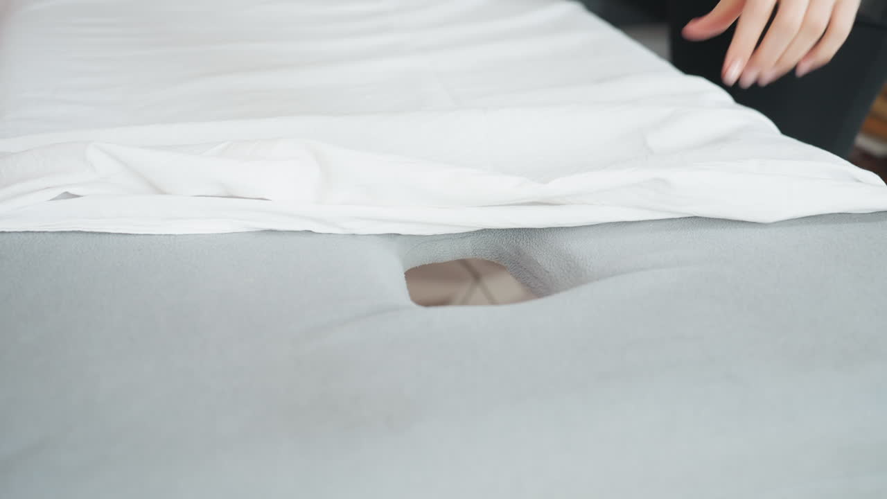 Woman carefully draping white bed sheet over light gray massage bed with face hole in clean interior setting, soft textures and calm lighting highlighting preparation of professional environment