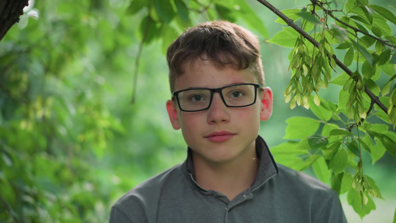 Close-up of young boy with pink lips wearing glasses and a grey top, blinking while focusing directly on the camera, a small insect flies in front of him, surrounded by blurred greenery