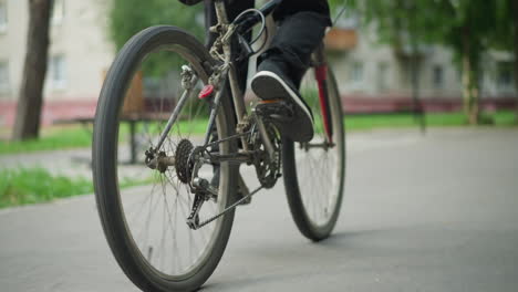 leg view of cyclist riding a bike on paved road, with a child riding a scooter in the opposite direction, background includes blurred view of benches, trees, greenery, and buildings