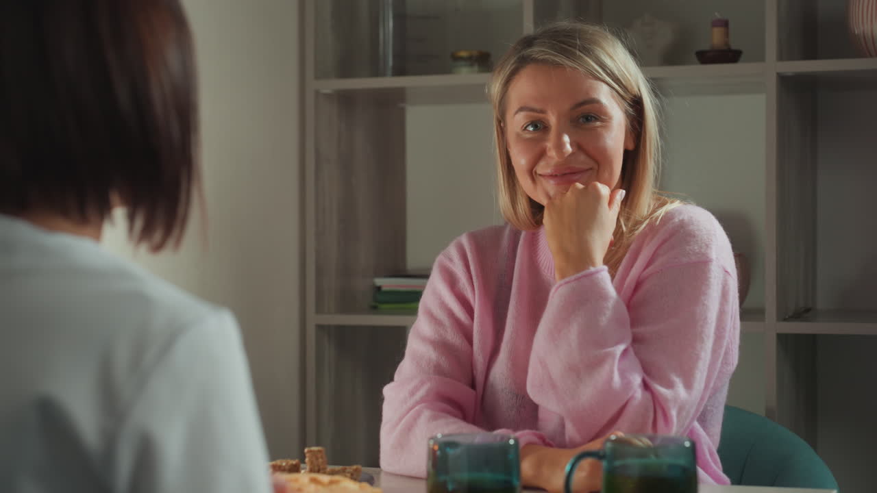 Donna bianca che si gode una sessione di microcorrente a casa, ambiente accogliente di cucina, mentore di bellezza che dimostra un dispositivo portatile, tazze sul tavolo, luce solare calda, conversazione amichevole, movimenti di massaggio delicati.