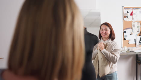 female tailor with cheerful expression removes beige blazer from black dress form while engaging in lighthearted conversation with partner inside bright studio surrounded by fashion sketches