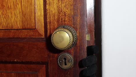 Un-Hombre-Con-Un-Guante-Negro-Abre-Lentamente-Una-Puerta-De-Madera-Girando-El-Pomo-De-La-Puerta-Y-Mostrando-Su-Mano-Y-Su-Rostro-Enmascarado