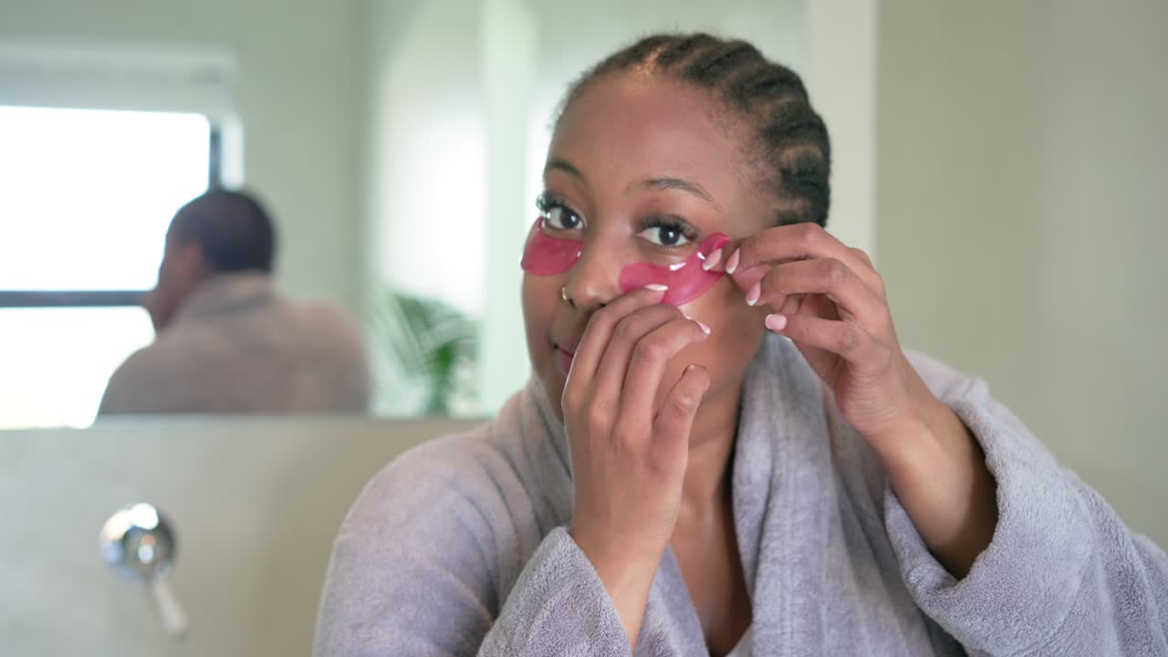 African American woman in robe retrieving, applying and peeling eye patches at mirror for refresh