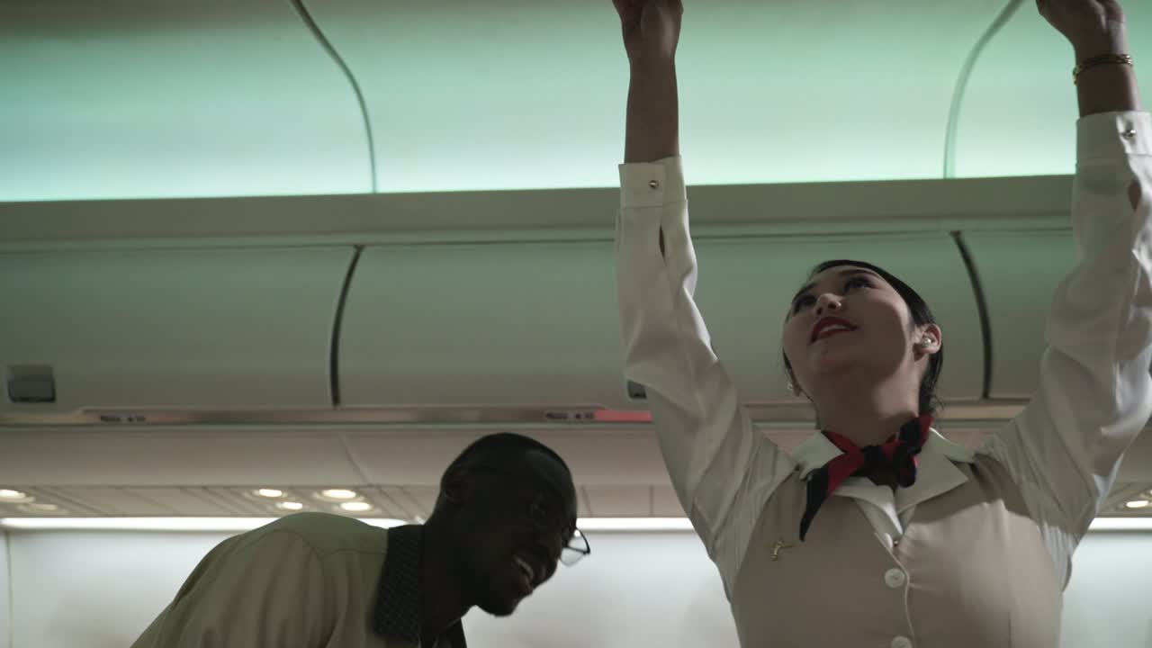 아시아 여성 승무원 또는 비행기 승무원이 유니폼을 입고 아프리카 남성 고객을 위해 도움의 서비스를 제공합니다. 객실 비행기의 상부 수하물 칸에 수하물을 보관하십시오.