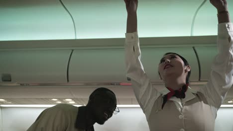 asian female flight attendant or airplane stewardess in uniform with help service for african man customer keep suitcase to overhead luggage compartment on cabin airplane.
