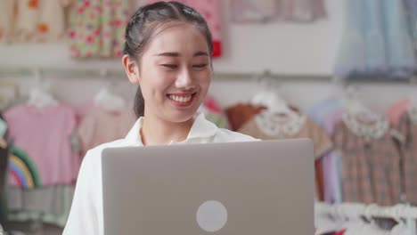 cerca de una mujer asiática dueña de una tienda de ropa para niños disfruta usando una computadora portátil, navegando por las redes sociales y haciendo marketing en línea después de comprobar las existencias en los rieles.