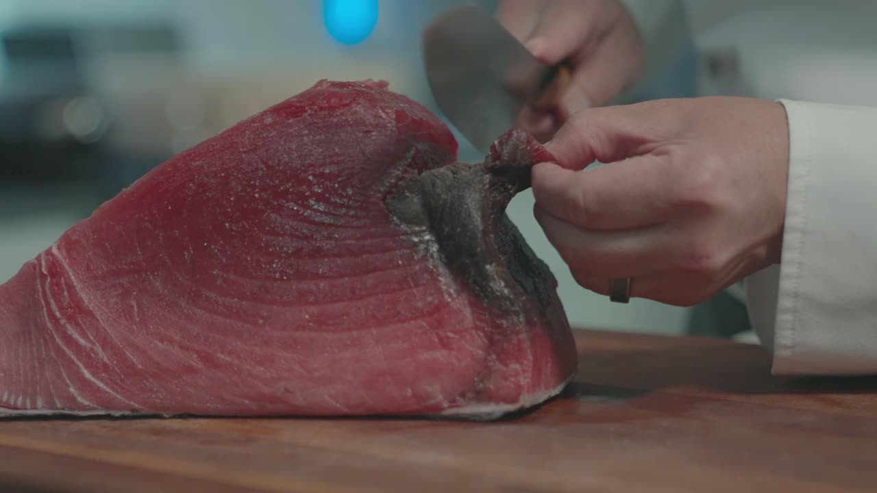 A static shot of a chef carefully removing the dark part of a tuna fish slab before preparing it, highlighting the attention to detail in the fish preparation process.