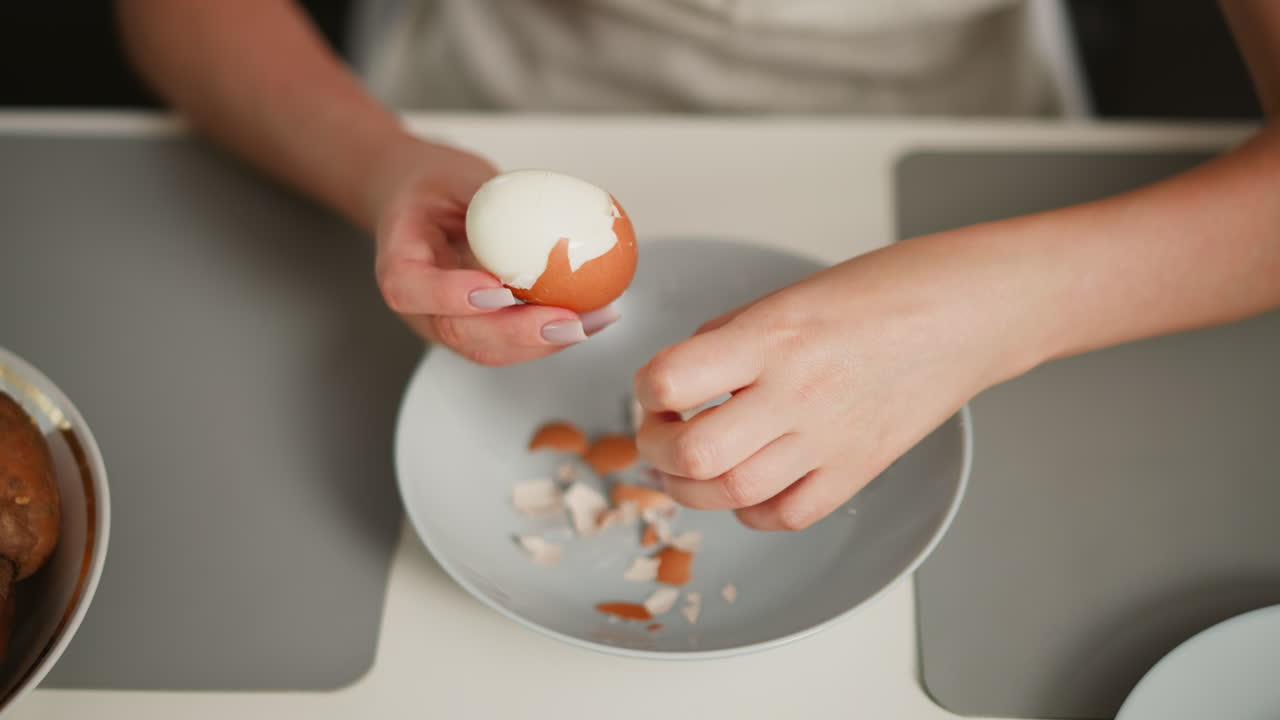 Overhead view of fair skinned person focused on peeling shell off boiled egg over bowl on table with scattered shell fragments, on placemat beside potatoes and empty dish