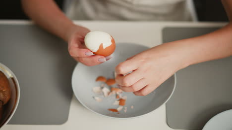 overhead view of fair skinned person focused on peeling shell off boiled egg over bowl on table with scattered shell fragments, on placemat beside potatoes and empty dish