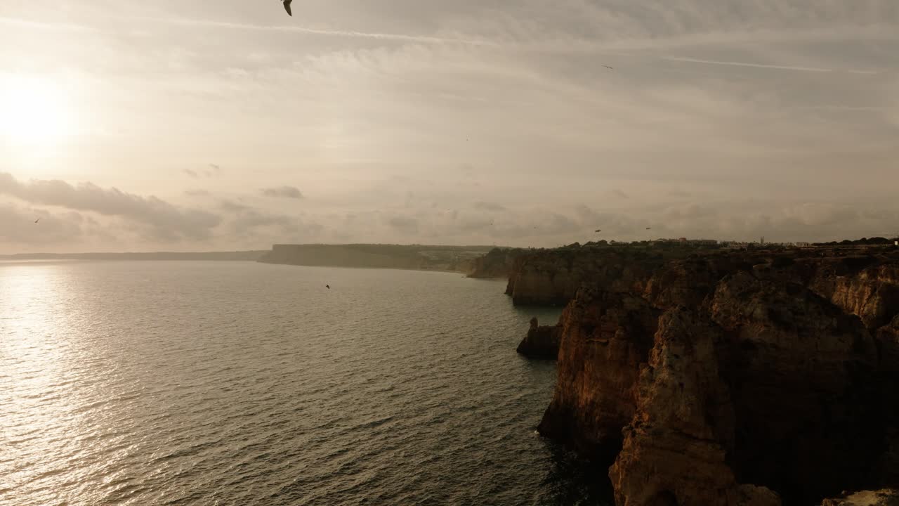 Vista aérea na hora dourada de falésias escarpadas e mar cintilante no Algarve, Portugal, com pássaros voando perto da costa.