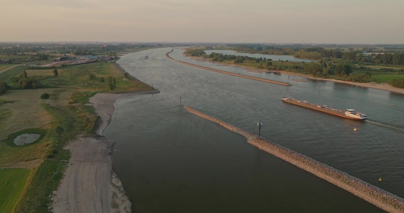 Un drone vola accanto a una nave mercantile che naviga attraverso un canale olandese al tramonto, rivelando campi verdi, dighe, piccole spiagge fluviali e il pacifico paesaggio rurale.