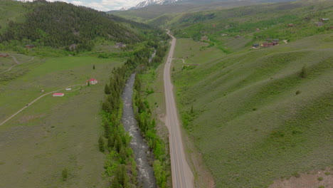 Aerial-view-of-beautiful-Colorado-road-and-mountain-range-with-snow-capped-peaks-on-a-sunny,-blue-sky-day-in-the-summer-with-green-fields,-trees,-and-mountain-homes