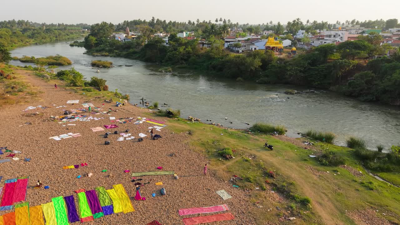 Vista aerea di un villaggio sul fiume dove le persone stendono panni colorati e lavano veicoli lungo la riva. Scena tradizionale di bucato rurale, fiume calmo e ambiente rurale pacifico