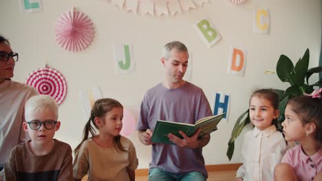 Un-Hombre-Con-Una-Camiseta-Morada-Y-Cabello-Gris-Y-Una-Niña-Con-Un-Peinado-Bob-Con-Una-Camisa-Blanca-Están-Sentados-En-Un-Círculo-Con-Niños-En-El-Suelo-Y-Leyendo-Un-Libro.-El-Hombre-Lee-Un-Libro-Para-Niños-En-Edad-Preescolar.