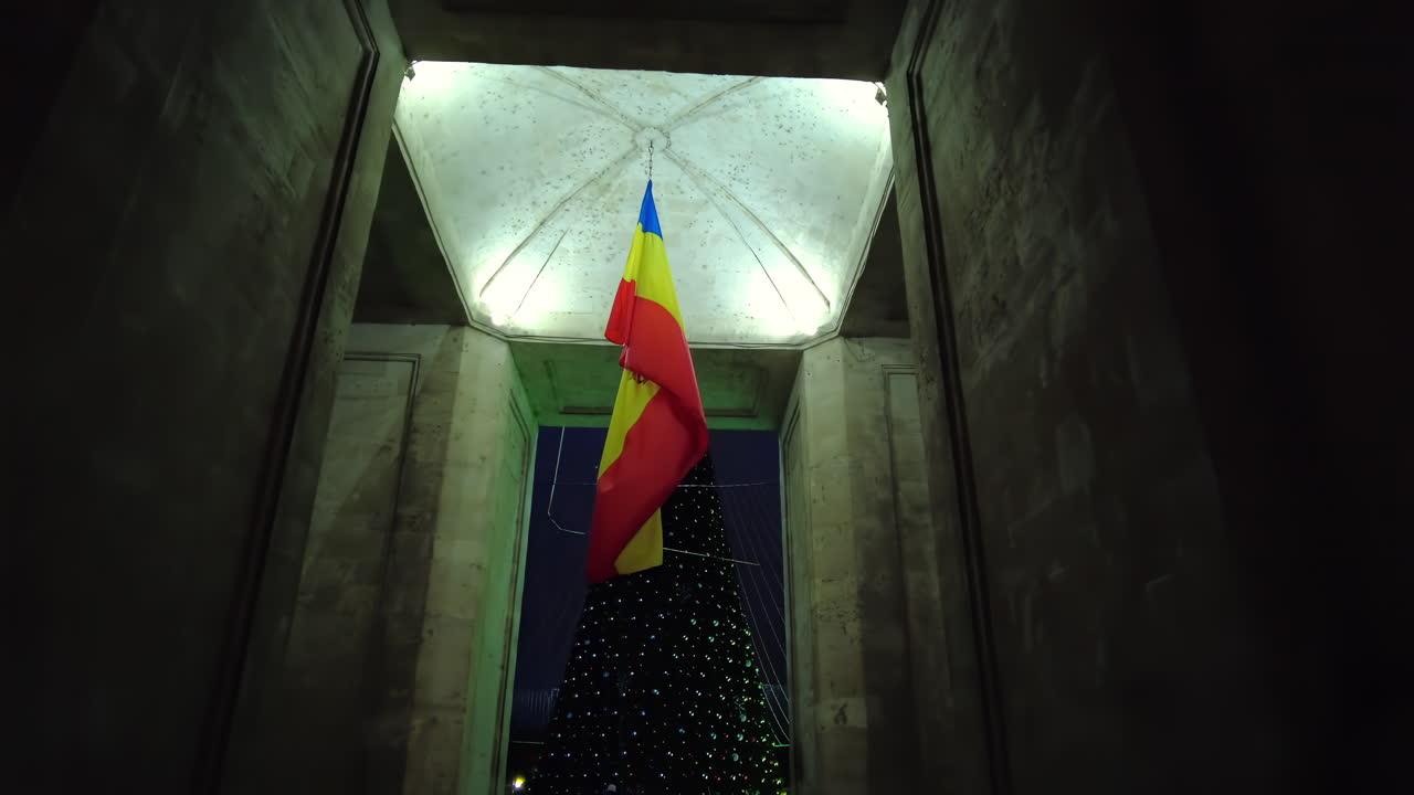 A large Romanian flag flutters under a grand structure as a beautifully decorated Christmas tree stands nearby. The scene captures a festive atmosphere in the evening