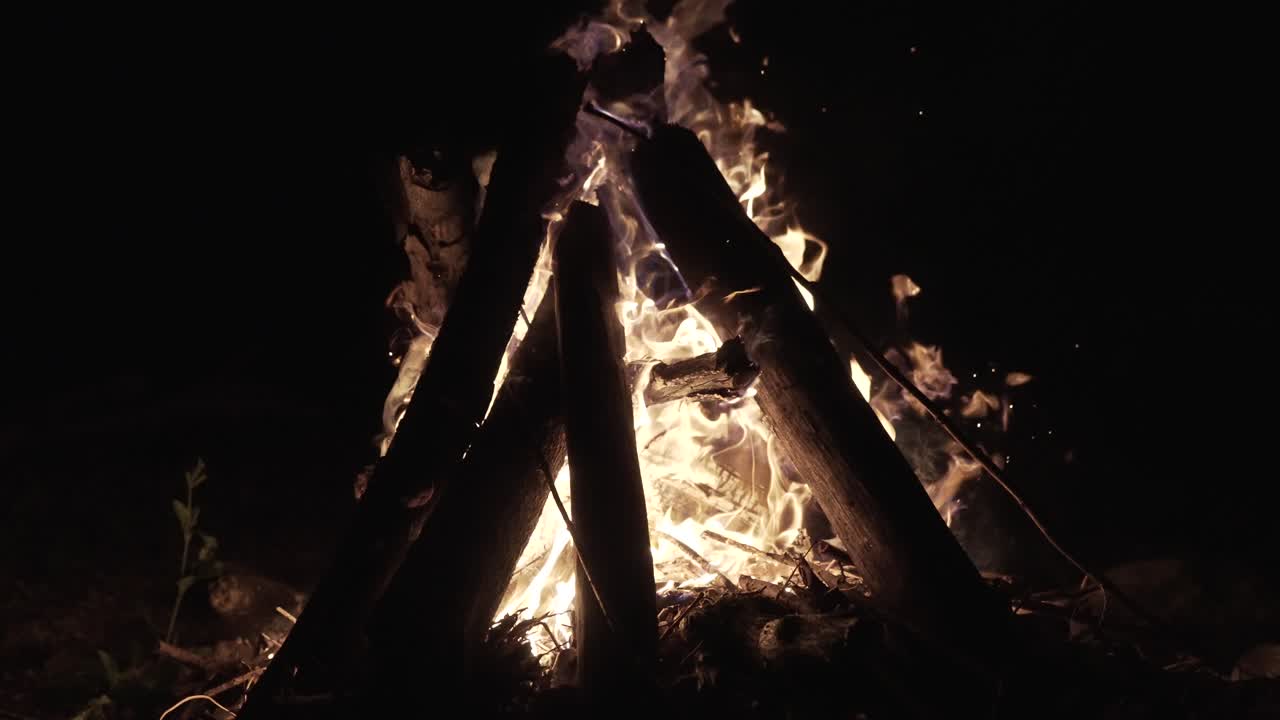 Fire burns wild, flames dance and flicker, wooden logs stacked in a teepee formation, sparks flying into the night air, creating warmth and light in the dark environment.