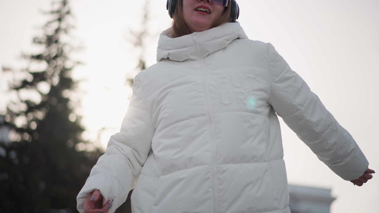 Creative dancer wearing white puffer jacket, beanie, black goggles and headphones turning around energetically with arms extended and fluid body gestures against pale winter sky in snowy urban plaza