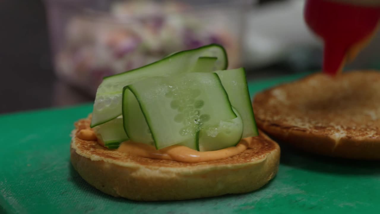 Close up, A chef prepares a burger by placing ribbons of fresh cucumber and swirls of spicy mayonnaise on the buns on a green chopping board