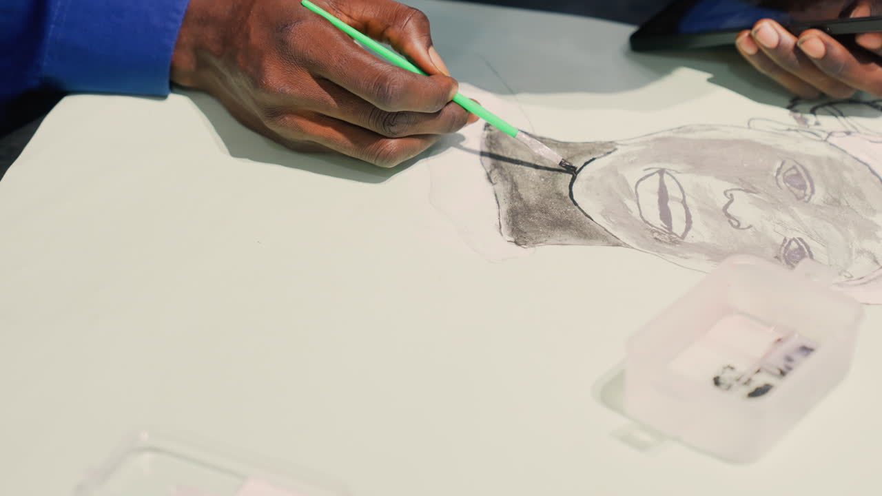 Closeup black portrait drawing in progress, artist hand stippling ink to define cheek and jawline with green stylus, plastic ink container and pencils at edge of white table, soft studio lighting