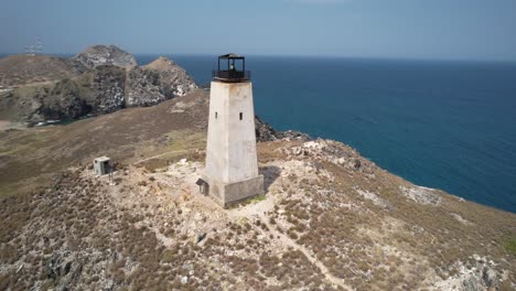 An-isolated-lighthouse-on-a-rocky-landscape-with-the-ocean-backdrop,-under-clear-skies,-aerial-view