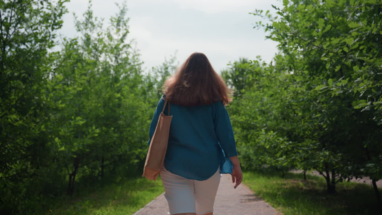 한 여성이 핸드백을 들고 공원 골목길을 따라 한가로이 거닐고 있고, 새는 하늘을 가로지르며 날아가고 있어요. 양쪽에는 나무가 있고, 여름의 푸르름이 가득하며, 평온한 분위기 속에서 천천히 걸으며, 마음을 챙기는 탈출, 자연 속 웰빙, 조용한 도시 속 오솔길을 느껴보세요.