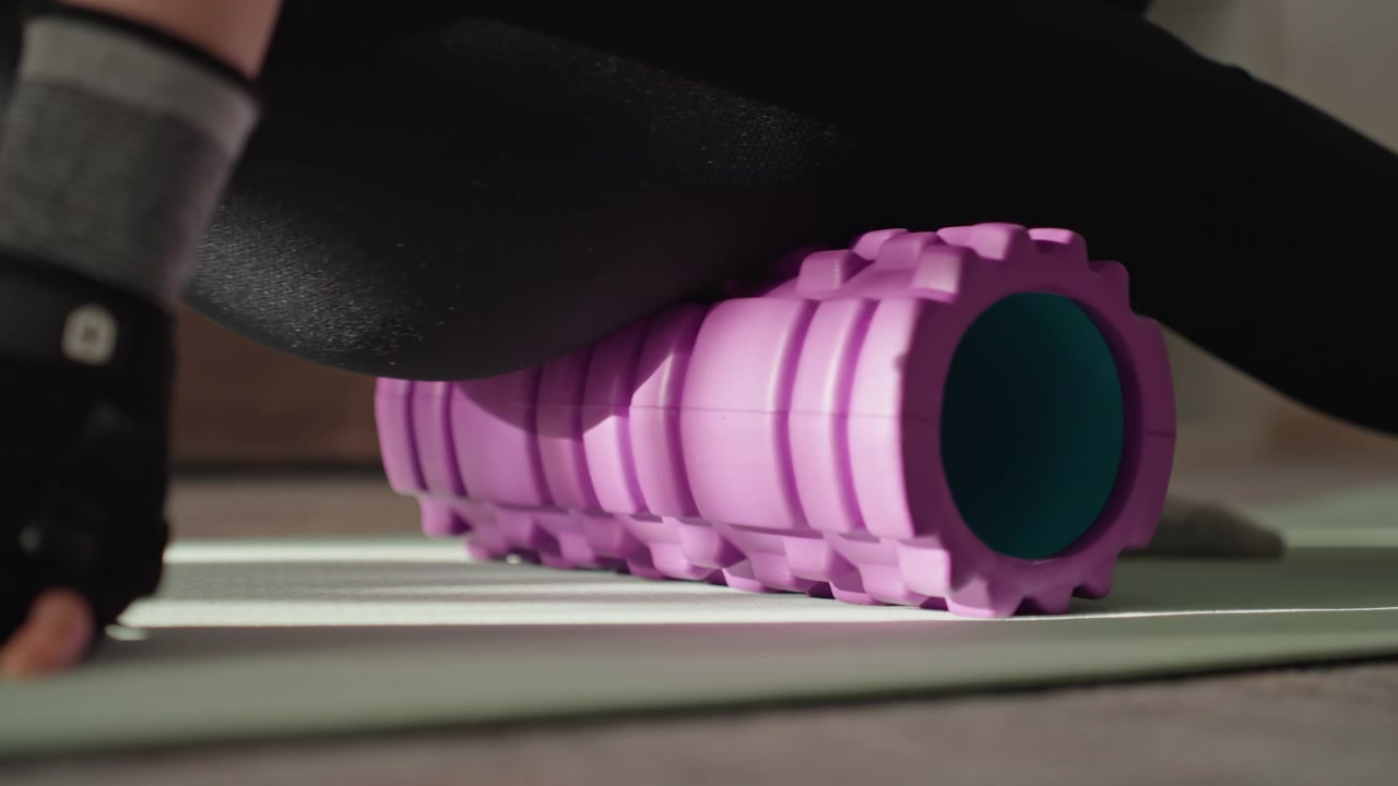 Home foam rolling session on mat, closeup of textured purple roller under thigh, person in black leggings and wrist grips applying pressure for muscle release and mobility, calm indoor lighting
