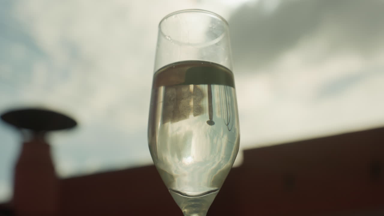 Bicchiere di champagne sul tetto illuminato dal sole che riflette lo skyline, lento primo piano di spumante in flûte con minuscole bollicine, silhouette sfocata del riscaldatore da esterno, cielo nuvoloso e malinconico, toni cinematografici caldi e superficiali