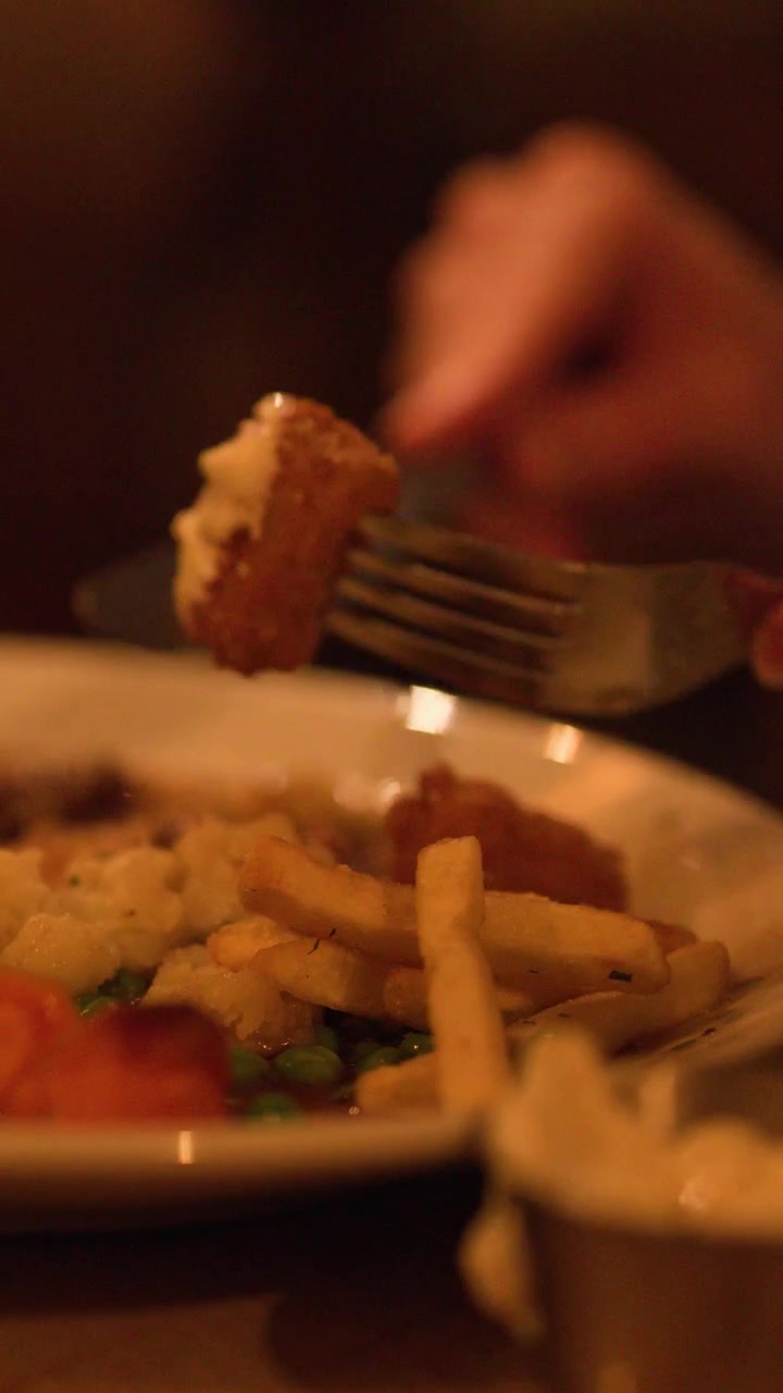 Mano che taglia e mangia un tortino di carne con patatine fritte, piselli e carote sotto una calda illuminazione ambientale da ristorante.