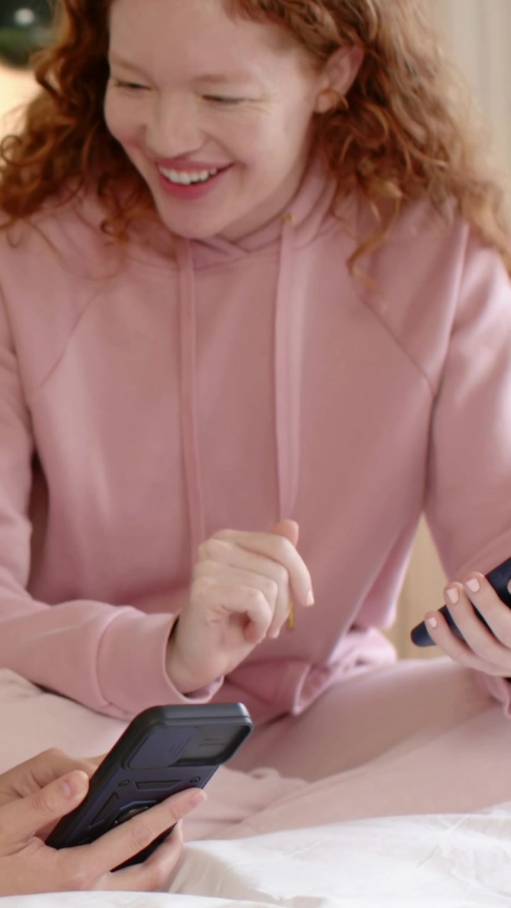 Vertical video: Smiling woman in pink hoodie glancing on bed after rugged phone appearing, reacting