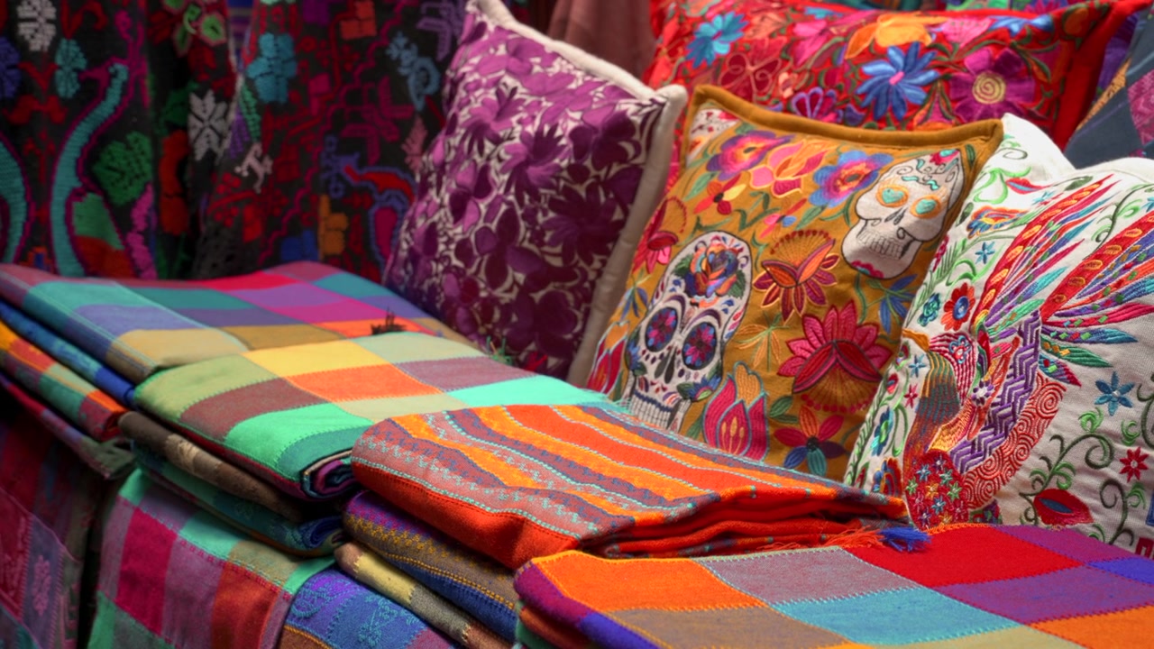 Static medium shot of folded colorful Mexican tablecloths and embroidered cushions with Day of the Dead skulls at a souvenir market.