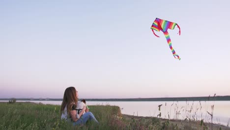 young happy woman and het little dog sitting with flying kite on a glade at sunset